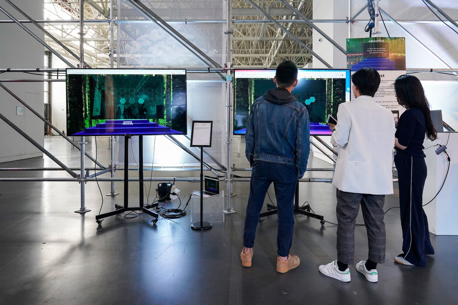 Visitors in front of two screens showing a forest and a purple stage, under a scaffold