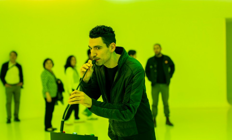 A person speaks into a microphone in a yellow-lit room while visitors watch
